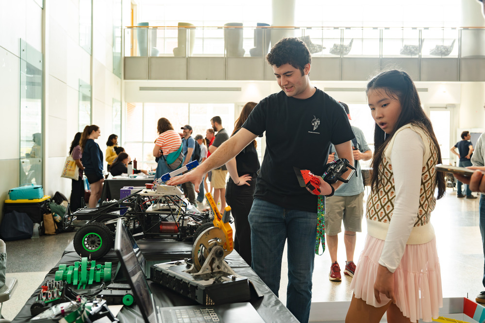 Visitors view student-built robotics and engineering projects on tables during a busy indoor science festival exhibition.